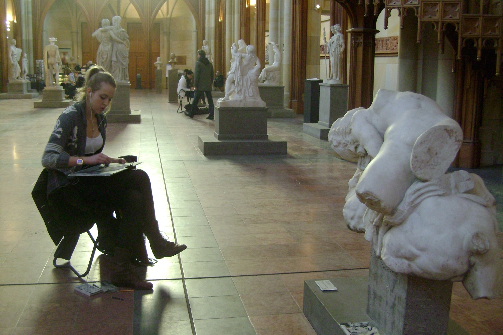 Junge Frau sitzt in einem Museum und zeichnet eine Plastik ab